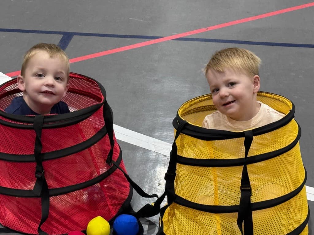 Two young children smiling while playing inside red and yellow mesh baskets on a gym floor